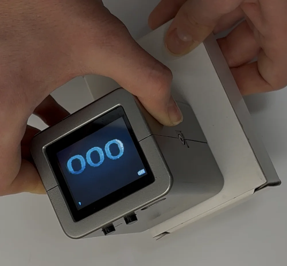 Hands holding the handheld imager against a white carton; the screen shows the glowing printed code 000